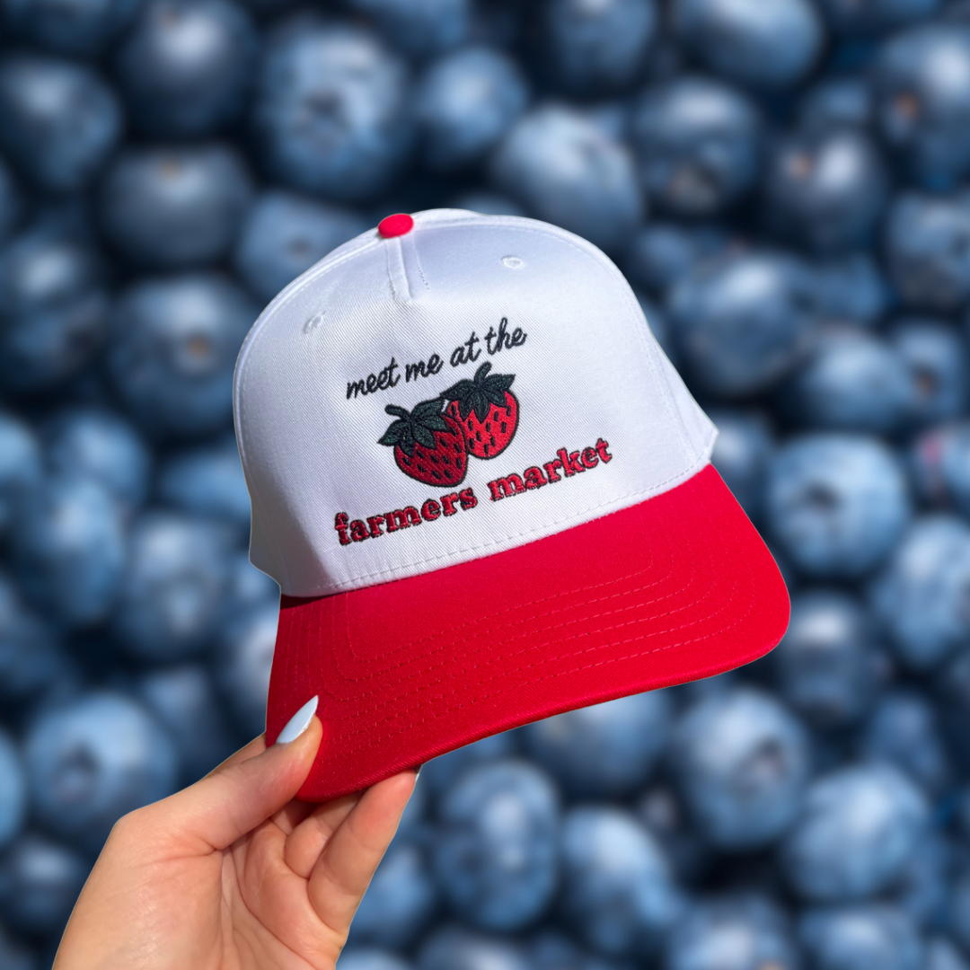 White and red cap with 'meet me at the farmers market' text held over a background of blueberries